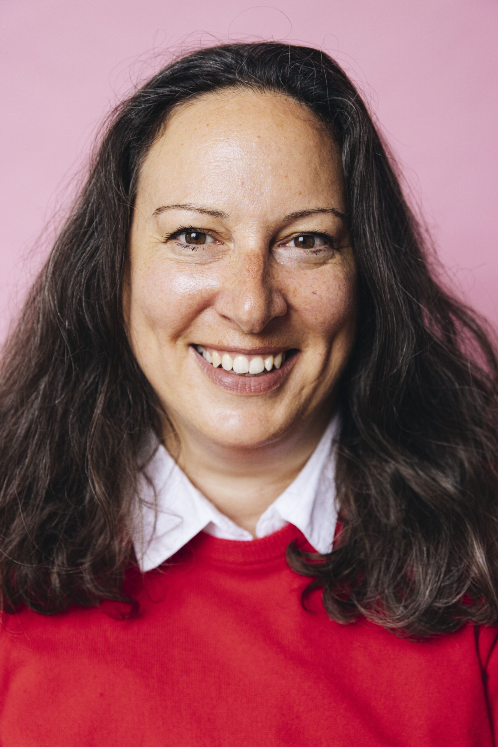 Lächelnde Frau mit dunklen Haaren und rotem Pullover vor rosa Hintergrund